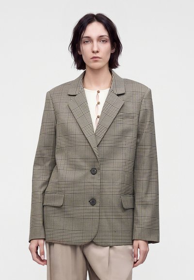 Jeune femme aux cheveux courts et foncés porte un blazer oversize à carreaux sur un haut crème et un pantalon beige, debout devant un fond uni.