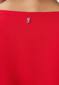 Prenda roja con una textura suave, que presenta un escote recto. Un acento de metal plateado está situado en la parte posterior.