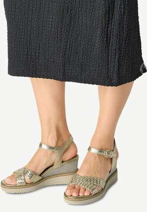 Metallisch goldene Keilsandalen mit einem geflochtenen Riemen-Design, verstellbarem Knöchelverschluss, strukturiertem Oberflächenmaterial und einer stabilen Sohle.