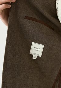Braun kariertes Blazer mit glatter Textur, ausgestattet mit einer braunen Akzenttasche und einem Etikett mit der Aufschrift "NEXT Tailoring, Relaxed Fit".