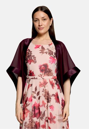 Frau trägt ein ärmelloses pinkfarbenes Kleid mit rotem und braunem Blumenmuster und einen transparenten, dunkelvioletten Schal über den Schultern.
