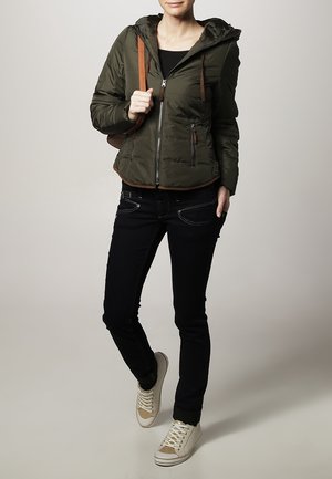 Vatteret jakke i olivengrøn med lynlåslukning foran og hætte, brune detaljer, kombineret med tætsiddende sorte jeans og cremefarvede sneakers.