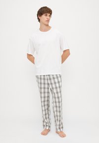 Vit kortärmad T-shirt i kombination med grå rutiga pyjamasbyxor med gröna och svarta detaljer. Modellen står barfota med händerna bakom ryggen.