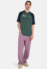 Camisa de manga corta verde y azul marino con un patrón pixelado, combinada con pantalones holgados de lavanda y zapatos beige. Ajuste casual y textura textil.