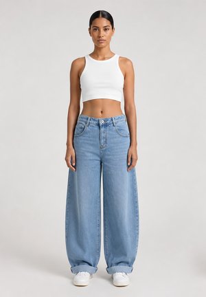 Kobieta stojąca, ubrana w biały bezrękawnik typu crop top, szerokie jasnoniebieskie dżinsy z podwiniętymi mankietami i białe trampki na jednolitym tle.