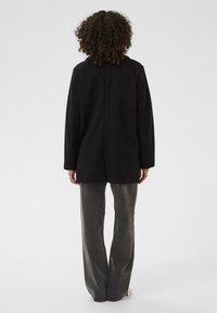 Čierny oversized blejzer s rovnou siluetou, jemnou Textúrou a čistým dizajnom. Nosený cez šedé, široké nohavice.