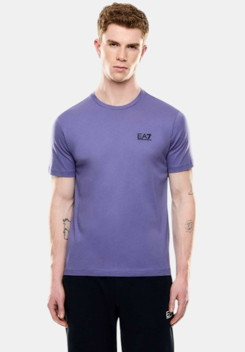 T-shirt di cotone viola con maniche corte, scollo tondo e logo EA7 nero sul lato sinistro del petto. Indossato con pantaloni scuri.