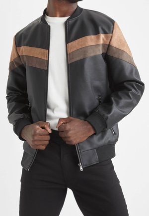 Mann trägt schwarze Bomberjacke mit braunen und beigen Wildlederstreifen, weißes Hemd darunter, schließt mit den Händen den Reißverschluss der Jacke.