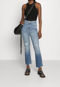 Top de tirantes negro acanalado, jeans de cintura alta azul claro desgastados con dobladillo sin terminar, y sandalias negras con tacones gruesos. Bolsa negra con correa trenzada.