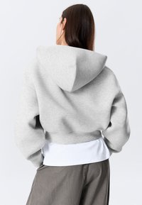 Kvinne med langt mørkt hår iført en lys grå cropped hettegenser over en hvit skjorte og grå bukser, sett bakfra mot en ensfarget bakgrunn.