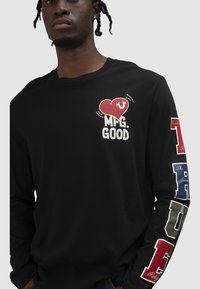 Mann trägt schwarzes Langarmshirt mit rotem Herz und dem Schriftzug "MFG. GOOD" auf der Brust sowie bunten Blockbuchstaben am rechten Ärmel.