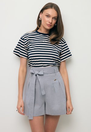 Kobieta o prostych brązowych włosach ubrana w granatowo-biały pasiasty t-shirt oraz jasnoszare szorty z wysokim stanem i przewiązanym paskiem, stojąca na białym tle.