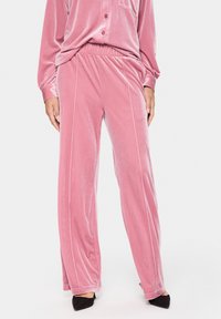 Sieviete, tērpusies rozā velveta platām biksēm un saskaņotu pogāto krekliņu, apvienojusi ar melniem šauriem zābakiem uz augstiem papēžiem pret baltu fonu.