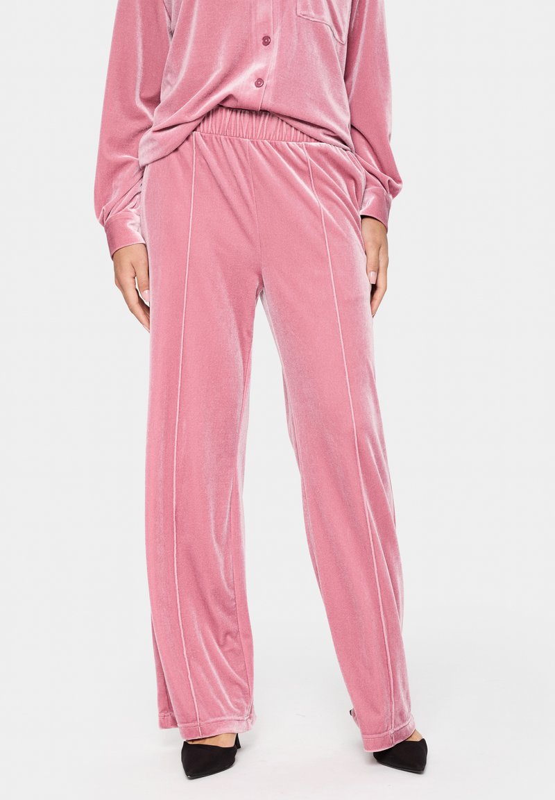 Sieviete, tērpusies rozā velveta platām biksēm un saskaņotu pogāto krekliņu, apvienojusi ar melniem šauriem zābakiem uz augstiem papēžiem pret baltu fonu.