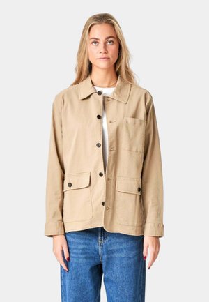 Beige bomuldsjakke med krave, frontknaplukning, to brystlommer og en løstsiddende pasform. Båret over en hvid top og blå jeans.
