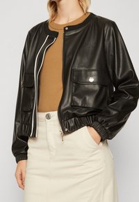 Sort imiteret læder bomberjakke med elastiske manchetter, lynlås lukning og to frontlommer. Har hvide detaljer og logo på brystet.