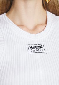 Gros plan d'une personne portant un haut côtelé blanc avec une étiquette "Moschino Jeans" noir et blanc sur la poitrine.