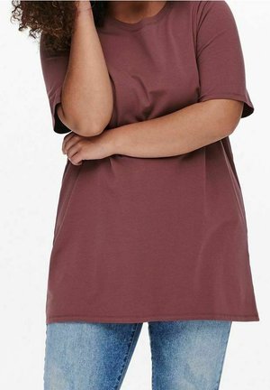 Frau mit lockigem Haar trägt ein lockeres dunkelrotes T-Shirt und hellblaue Jeans, mit einem Arm verschränkt und der anderen Hand am Kopf.