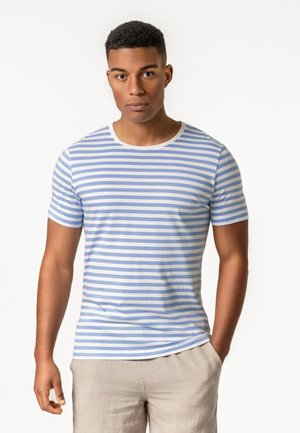 Junger Mann steht, trägt ein hellblau-weiß gestreiftes kurzärmeliges Hemd und beige Hose, blickt geradeaus.