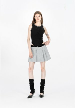 Joven mujer con top negro sin mangas, falda plisada gris con cinturilla negra, calentadores de piernas negros fruncidos y zapatos de tacón negro, de pie.