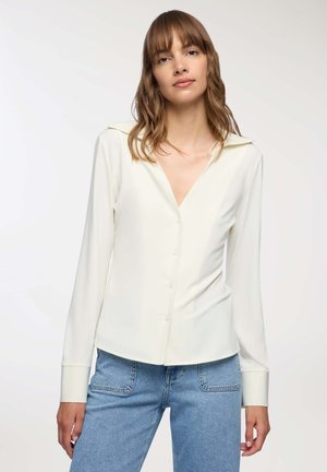 Donna che indossa una camicia a maniche lunghe di colore crema con colletto ampio e jeans azzurri, in piedi contro uno sfondo chiaro.