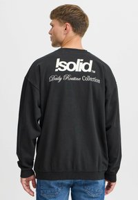 Czarna bluza z luźnym krojem, z dużym białym logo i napisem na plecach. Rękawy i dół wykończone ściągaczem, miękka faktura, casualowy design.