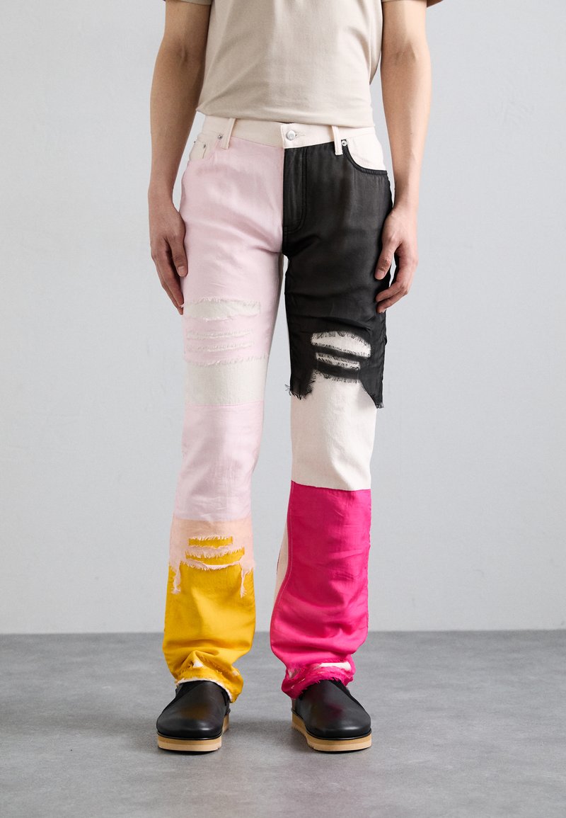 Jeans di denim colorati caratterizzati da un design a patchwork con sezioni rosa, nere, bianche e gialle; dettagli strappati sulle ginocchia.