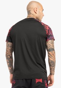 Kortærmet skjorte med sort krop og røde/lilla mønstrede ærmer. Lavet af glat stof. Omfatter TAPOUT-logoet i bunden.