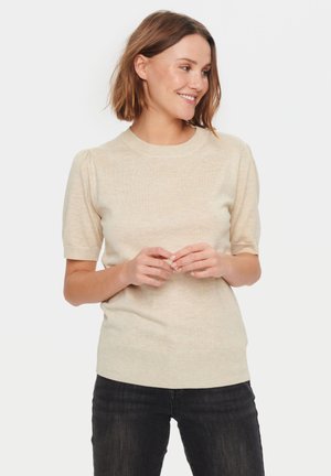 Maglione a maniche corte beige in maglia con scollatura rotonda e sottili spalle a sbuffo. Il tessuto ha una finitura morbida e texture. Indossato con jeans scuri.