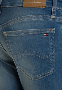 Vaqueros de mezclilla azules con una vista en primer plano del bolsillo trasero, que presenta costuras beige y un pequeño logotipo rojo, blanco y azul.