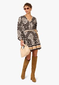 Schwarz-weiß gemustertes Kleid mit langen Ärmeln und Taillengürtel, kombiniert mit kniehohen hellbraunen Wildlederstiefeln und einer beigen Mini-Handtasche.