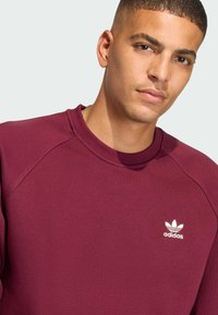 Maroon sweatshirt with a crew neck and raglan sleeves. Features a small white Adidas logo on the chest. Fabric appears soft and textured.