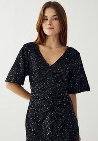 Juoda blizgučių suknelė su V formos iškirpte, trumpomis rankovėmis ir surinkimo detalėmis aplink liemenį, kuri suteikia tekstūruotą, spindinčią paviršių.