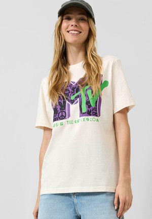 Jeune femme portant un t-shirt graphique MTV de couleur claire, un jean bleu et une casquette de baseball sombre, souriant sur un fond uni.