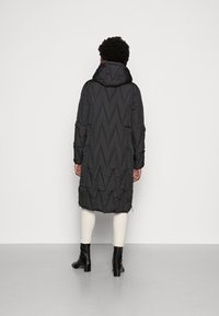 Manteau matelassé noir avec capuche, présentant un design en losanges, manches longues et fermetures éclair latérales. Porté sur des leggings clairs et des bottes.