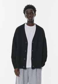 Un cardigan tricotat negru, cu margini ribbate și închidere cu șase nasturi, purtat peste o cămașă albă, asociat cu pantaloni de trening gri.