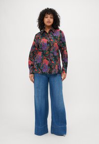 Blusa nera a fantasia floreale con swirl colorati, chiusura frontale con bottoni e maniche lunghe, abbinata a jeans in denim blu a gamba larga.