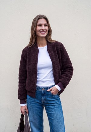Lächelnde Frau, die eine dunkelviolette Fleecejacke, ein weißes Hemd und blaue Jeans trägt und eine braune Tasche in einer Hand vor einer schlichten Wand hält.