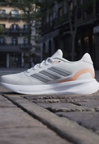 Chaussure de sport blanche avec des rayures grises et une touche pêche sur une rue pavée en milieu urbain, vue latérale montrant la semelle et les lacets.