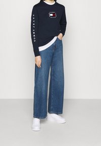 Laivastonsininen pitkähihaisuus paita valkoisilla yksityiskohdilla ja rintaloggolla, yhdistettynä leveisiin sinisiin farkkuihin. Mallilla on valkoiset tennarit.