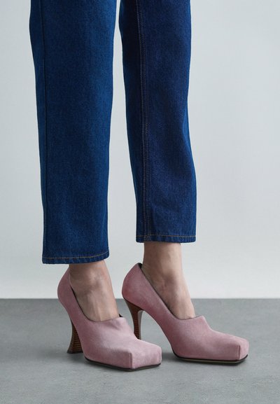 Ροζ σουέτ ψηλοτάκουνα παπούτσια με τετράγωνη μύτη και καμπύλη φτέρνα, διαθέτοντας ξύλινη φτέρνα με υφή και λείο επάνω μέρος. Φορεμένα με μπλε τζιν παντελόνι.