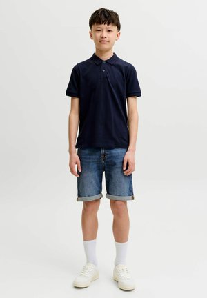 Gutt står, iført marineblå poloskjorte, oppbrettede denimshorts, hvite sokker og hvite joggesko mot ensfarget lys bakgrunn.