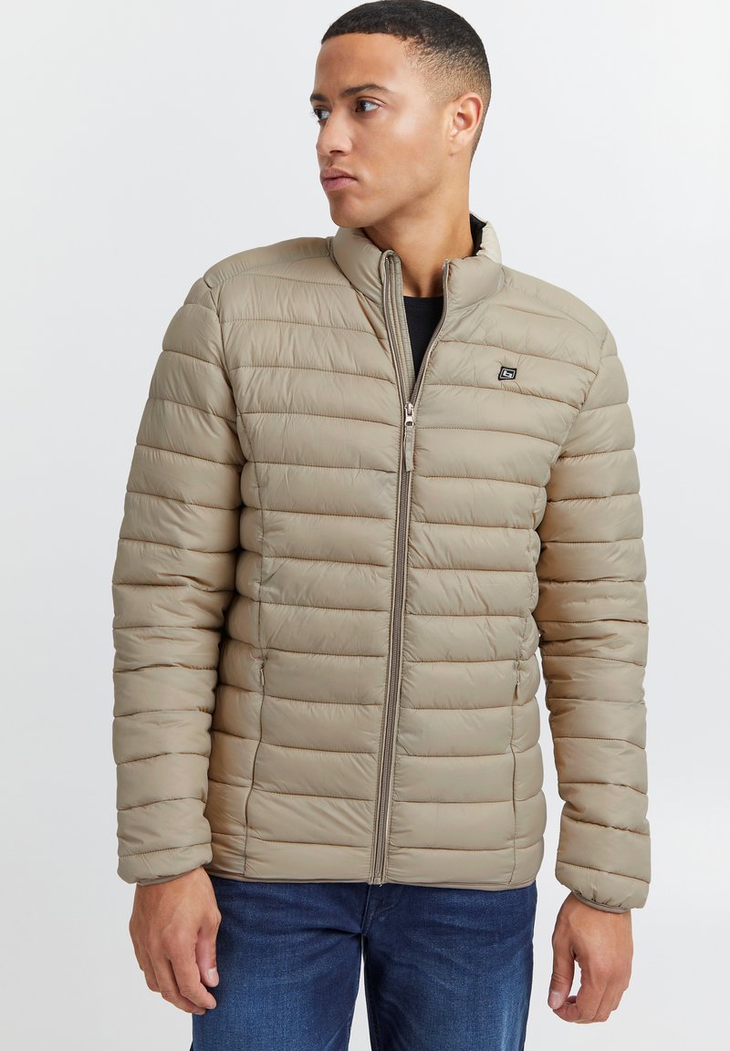 Chaqueta acolchada beige con costuras horizontales, cuello alto, cremallera completa y detalle de logo en el pecho. Llevada sobre una camisa negra.