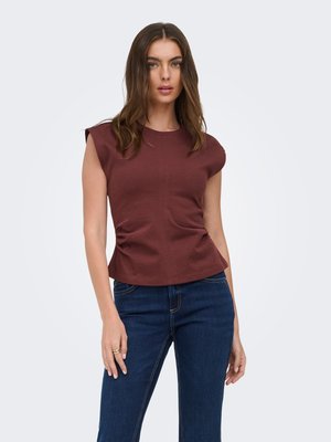 Kvinde med langt brunt hår iført en ærmeløs mørkerød top og blå jeans, stående mod en ensfarvet lys baggrund.