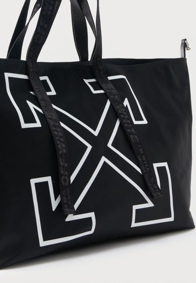 Μαύρη τσάντα tote με μεγάλο λευκό γραφικό διχαλωτού βέλους και μαύρα λουριά με διακριτικό λογότυπο.