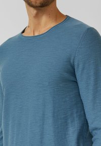 Mann trägt einen blauen Langarmpullover mit Rundhalsausschnitt, Oberkörper und Hals vor einem einfarbigen Hintergrund sichtbar.