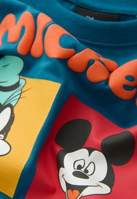 Teal bomulds t-shirt med levende orange tekst og farverige tegneseriefigur designs, herunder Mickey Mouse og Goofy, mod en kontrasterende baggrund.
