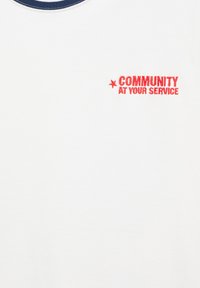 T-shirt blanc avec détail col bleu marine et texte rouge sur la poitrine indiquant "COMMUNITY AT YOUR SERVICE" accompagné d'une icône d'étoile rouge.