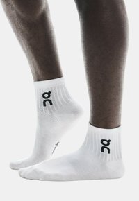 Calcetines deportivos blancos con textura acanalada, que presentan un logo negro en el lateral y áreas reforzadas en el talón y los dedos para mayor durabilidad.