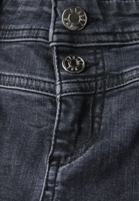 Mörka jeans i denim med en närbild av två metallknappar, synlig söm och en framficka med en mjuk, texturerad finish.